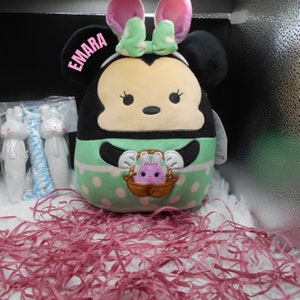 May include: A plush toy of a cartoon mouse wearing a green and white dress and a green bow. The toy has large black eyes and a pink nose. The toy is holding a basket with a pink bunny inside. The toy is sitting on a pink and white shredded paper.