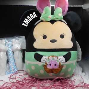 May include: A plush toy of a cartoon character with black ears, a green and white polka dot dress, and a basket with a pink bunny inside. The toy has a name tag that says "EMARA".