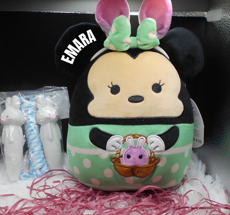 May include: A plush toy of a cartoon character with black ears, a green and white polka dot dress, and a basket with a pink bunny inside. The toy has a name tag that says "EMARA".