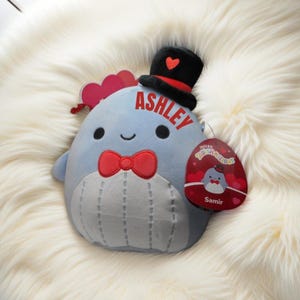 May include: A blue plush toy with a red bow tie and a black top hat with a red heart. The toy is named "Ashley" and has a tag that says "Original Squishmallows" with a smaller plush toy named "Samir".