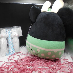 May include: A black and green plush toy with white bunny ears and polka dots. The toy is sitting in a box with pink shredded paper.