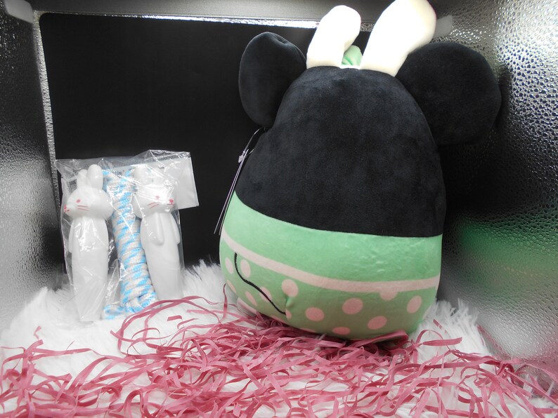 May include: A black and green plush toy with white bunny ears and polka dots. The toy is sitting in a box with pink shredded paper.