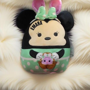 May include: A green, white, and black plush toy shaped like a cartoon mouse with bunny ears. The toy has a pink bow and the name "EMARA" embroidered on the front.