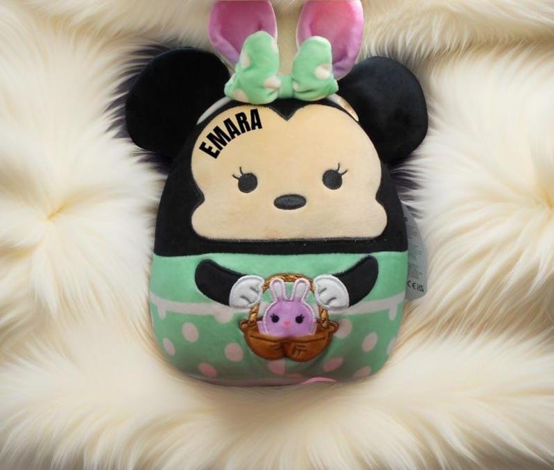 May include: A green, white, and black plush toy shaped like a cartoon mouse with bunny ears. The toy has a pink bow and the name "EMARA" embroidered on the front.