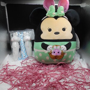 May include: A green and white plush toy shaped like a mouse with bunny ears. The toy is holding a basket with a small purple bunny inside. The toy is sitting on pink shredded paper in a box.