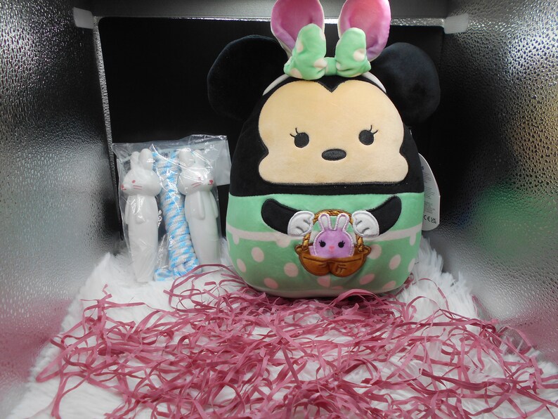 May include: A green and white plush toy shaped like a mouse with bunny ears. The toy is holding a basket with a small purple bunny inside. The toy is sitting on pink shredded paper in a box.