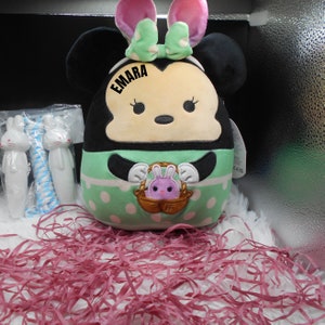 May include: A green and white plush toy with black ears and a pink bow. The toy has a face with black eyes and a pink nose. The toy is holding a basket with a pink bunny inside. The toy has the name "EMARA" written on it.