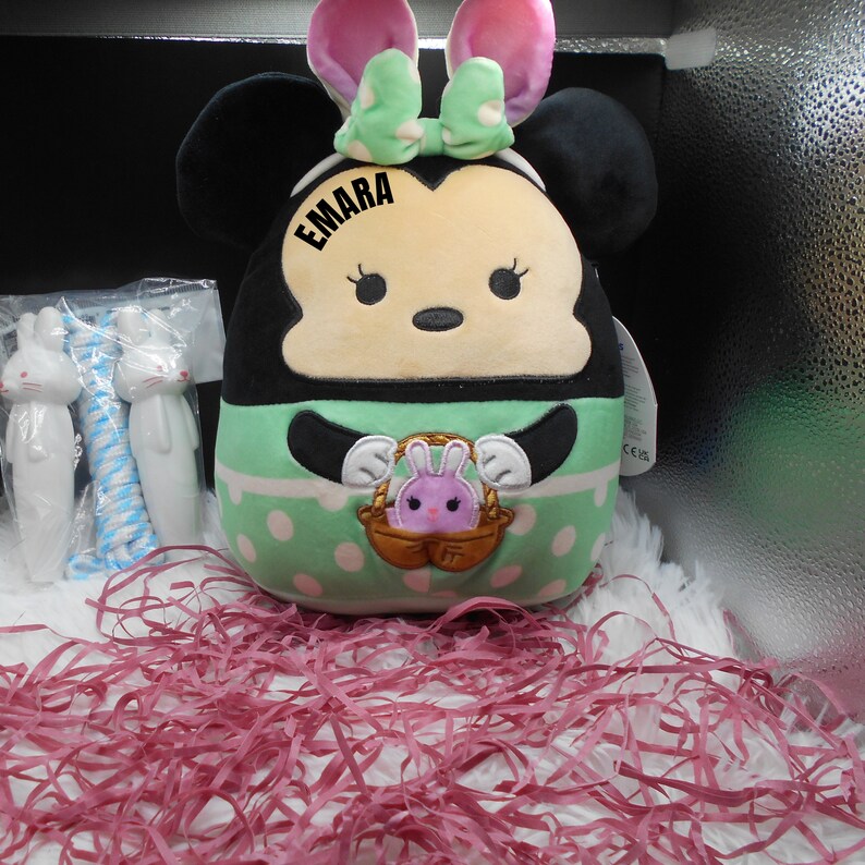 May include: A green and white plush toy with black ears and a pink bow. The toy has a face with black eyes and a pink nose. The toy is holding a basket with a pink bunny inside. The toy has the name "EMARA" written on it.