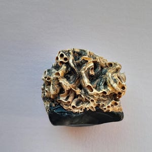 Könnte beinhalten: Eine strukturierte Skulptur mit dunkler Basis und detailliertem, porösem Oberteil. Die Oberseite weist eine Mischung aus Beige-, Braun- und Off-White-Farben auf, wodurch ein natürliches, organisches Aussehen entsteht. Die Basis der Skulptur ist dunkel und glänzend.
