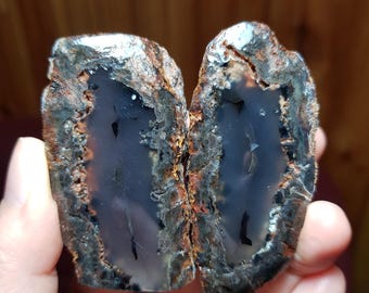 Puede incluir: Dos mitades de una geoda con un interior de ágata gris y un exterior marrón oscuro, negro y gris.