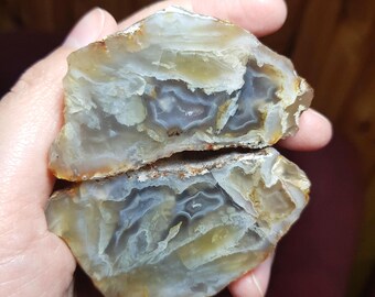May include: Two halves of a polished agate stone, showcasing intricate patterns of gray, white, and yellow hues. The stone's interior reveals layered formations, with a rough, brown exterior. The cut surfaces display a natural, organic aesthetic.