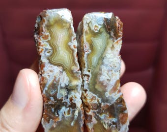 May include: Two polished agate stones in shades of brown, white, yellow, and green. The stones have a rough edge and a smooth, polished face. The stones are held in a hand.