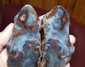 Op de afbeelding: Twee helften van een gepolijste agaatsteen, met ingewikkelde patronen van blauw, rood en bruin. Het oppervlak van de steen is glad en onthult kleurlagen en natuurlijke formaties. De helften worden bij elkaar gehouden.