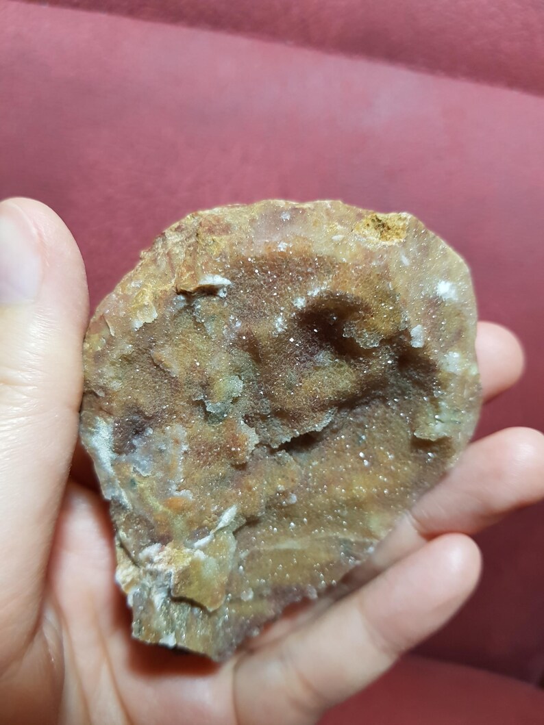 May include: A rough, irregularly shaped rock formation with a mix of brown, tan, and white crystalline structures. The surface has a textured, granular appearance, with small sparkling crystals scattered throughout. The rock is held in a hand against a red background.