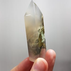 May include: A clear, pointed crystal with a frosted appearance. The crystal has a natural, earthy aesthetic, with areas of green and brown inclusions. The crystal is held in a hand against a plain background.