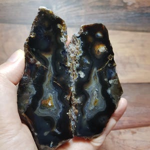 Può includere: Due metà di una pietra d'agata lucidata, che mostra un motivo scuro e stratificato con bande di nero, marrone e bianco. La pietra naturale ha un bordo ruvido e irregolare e una superficie lucida e liscia. L'agata è tenuta in una mano.