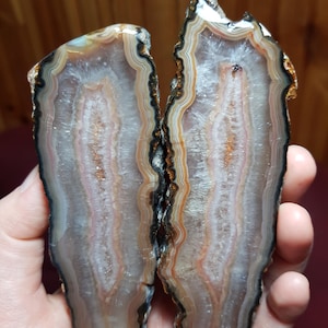 May include: Two halves of a large agate geode with a white, pink, and brown banded pattern. The agate has a black outer edge and is cut in a long, thin shape.