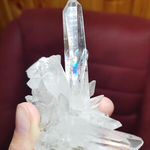 May include: A cluster of clear quartz crystals with sharp, pointed terminations. The crystals are translucent, with some displaying iridescent rainbow reflections. The quartz is held in a hand, showcasing its natural, raw form. The overall appearance is of a natural mineral specimen.
