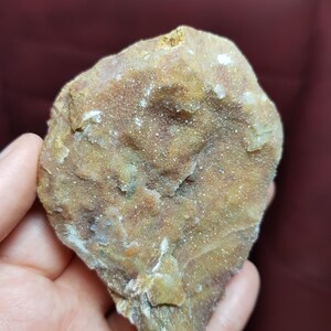 May include: A rough, irregularly shaped rock with a mix of tan, brown, and white colors. The surface has a textured, crystalline appearance, with small, sparkling crystals visible throughout. The rock is held in a hand, showcasing its natural, earthy tones.
