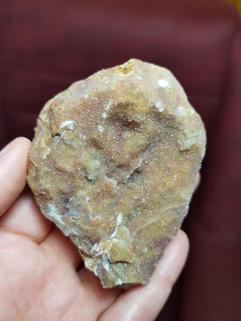 May include: A rough, irregularly shaped rock with a mix of tan, brown, and white colors. The surface has a textured, crystalline appearance, with small, sparkling crystals visible throughout. The rock is held in a hand, showcasing its natural, earthy tones.
