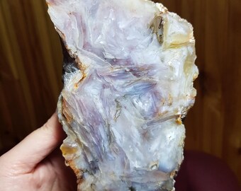 May include: A large, raw gemstone with a layered appearance. The stone features a mix of colors, including white, lavender, and brown. The rough texture and natural shape of the stone are visible. The gemstone is held in a hand.