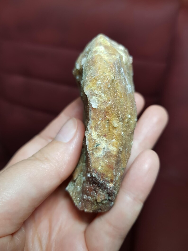 May include: A rough, natural crystal formation with a light brown and tan color. The crystal has a textured surface and is held in a hand.