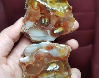 May include: Two rough-cut agate stones in shades of orange, white, yellow, and brown. The stones have a natural, organic shape and texture, with visible layers and patterns. The agate pieces are held in a hand.