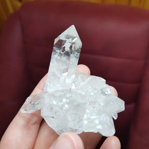 May include: A clear quartz crystal cluster with multiple points and facets. The crystal is held in a hand, showcasing its natural, translucent appearance. The quartz has a crystalline structure, reflecting light.