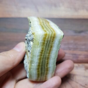 May include: A natural stone specimen with layers of translucent, pale green and brown bands. The stone has a rough exterior and a polished, layered interior. The overall appearance is earthy and organic, suitable for collectors or decorative use.