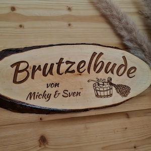 Op de afbeelding: Ovaal houten bord met de gegraveerde tekst "Brutzelbude von Micky & Sven". Het bord bevat een illustratie van een saunatemmer en -kwast. Het bord heeft een donkerbruine schorsrand en is gemaakt van lichtgekleurd hout.