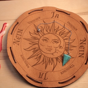 Può includere: Un tabellone di pendolo in pelle marrone con un design di sole e luna. Il tabellone ha le parole "Ja", "Nein" e "Unsicher" stampate su di esso. Un pendolo turchese è appeso a una catena d'argento.
