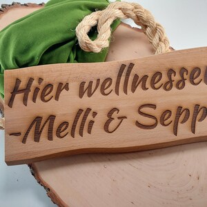 Könnte beinhalten: Ein Holzschild mit dem Text "Hier wellnessen - Melli & Seppi -" darauf. Das Schild hängt an einem Seil und liegt auf einer Holzoberfläche.