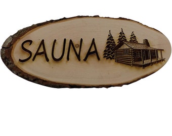 Puede incluir: Un letrero de madera con la palabra "SAUNA" tallada en él. El letrero tiene forma ovalada y tiene un aspecto rústico y natural. Una pequeña cabaña con árboles está tallada en la madera en el lado derecho del letrero.