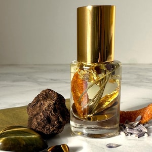 Pode incluir: Uma garrafa de vidro com tampa dourada cheia de um líquido transparente e flocos dourados, casca de laranja e lavanda seca. A garrafa está sobre uma superfície de mármore branco com uma rocha marrom, uma folha verde e várias pedras de olho de tigre.