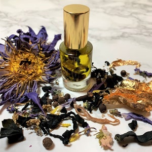 Puede incluir: Una pequeña botella de vidrio con tapa dorada llena de un líquido amarillo y hierbas secas. La botella está rodeada de flores secas, hierbas y pan de oro.