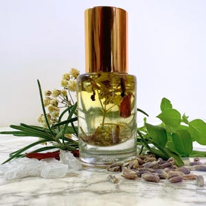 May include: A clear glass bottle with a gold cap filled with a yellow liquid and botanicals. The bottle is surrounded by sprigs of rosemary, lavender buds, and other herbs. The background is a white marble surface.