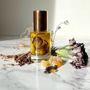 Puede incluir: Un frasco de perfume de vidrio transparente con tapa color cobre, lleno de un líquido amarillo y una flor seca. El frasco está rodeado de hierbas secas, trozos de resina de color ámbar y una flor morada parcialmente seca. Fondo de mármol.