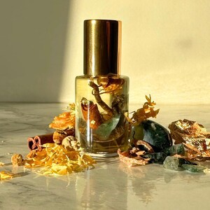 Puede incluir: Una botella de vidrio transparente con tapa dorada, llena de un líquido claro y elementos botánicos. La botella está rodeada de flores secas, palitos de canela y piedras preciosas. La estética general es natural y terrosa.