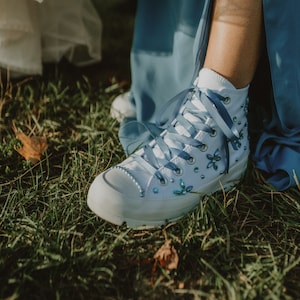 Puede incluir: Zapatillas blancas de lona de caña alta con cordones de satén azul y adornos de pedrería azul. Las zapatillas las lleva una persona que lleva un vestido azul.