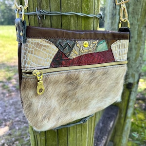 May include: A crossbody bag featuring a lower section of tan and white cowhide fur and an upper section of multi-colored, textured leather patches in cream, red, and green. The bag includes a gold-tone zipper, metal hardware, and a dark brown strap.