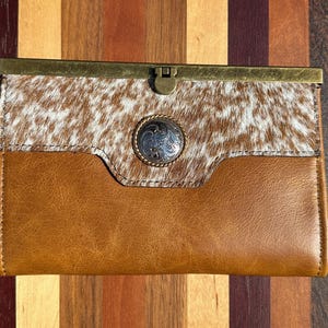 May include: A rectangular clutch wallet featuring a brown leather lower section and a white and brown cowhide upper section. The wallet has a brass-toned metal frame closure and a circular silver-toned concho ornament centered on the front.