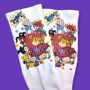 May include: A pair of white socks with a colorful graphic print of the Rugrats characters. The socks feature the Rugrats logo and the characters Tommy Pickles, Chuckie Finster, Phil and Lil DeVille, Angelica Pickles, and Susie Carmichael.
