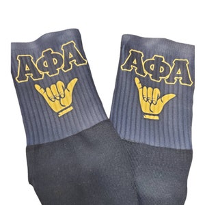 May include: A pair of black socks with a gold "AΦA" logo and a gold hand gesture on each sock.