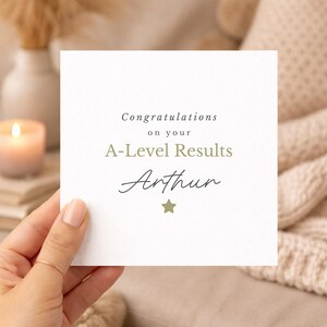 May include: A square white greeting card with black and olive green text that reads Congratulations on your A-Level Results Arthur, featuring a small olive green star icon centered at the bottom of the card design.