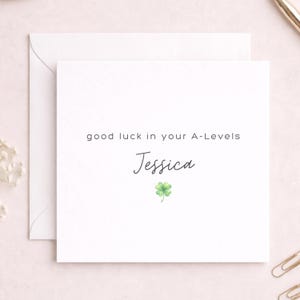 May include: A square white card with the message "good luck in your A-Levels" and the name "Jessica" in a script font. A small green four-leaf clover is at the bottom. The card is in a white envelope.