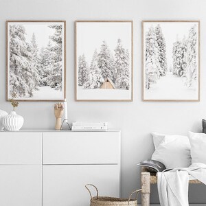 May include: Three framed wall art prints featuring black and white winter forest scenes. The center print displays a small wooden A-frame cabin nestled among snow-covered pine trees. Each print is encased in a light-toned wooden frame.