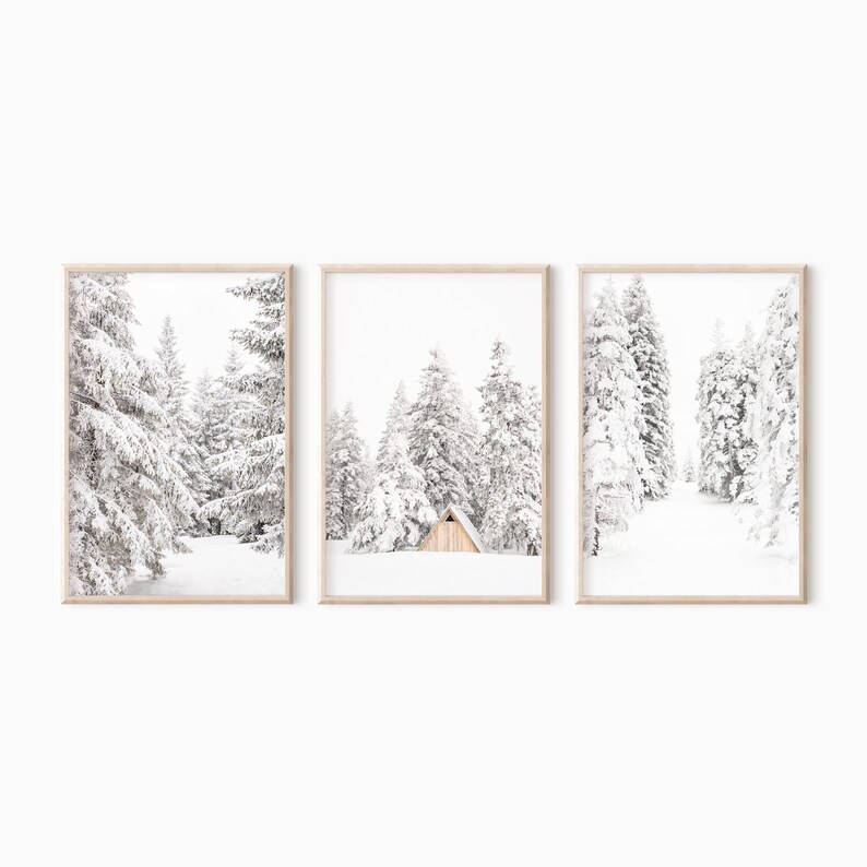 May include: A triptych of three vertical framed prints featuring a winter landscape. Each print displays snow-covered evergreen trees and a white, snowy ground. The center print includes a small, light-colored wooden A-frame cabin nestled in the trees.