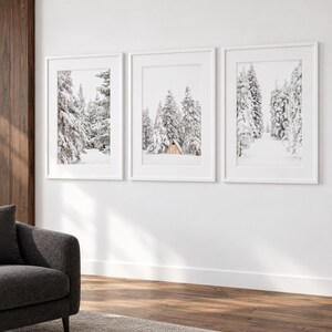 May include: Three vertical framed wall art prints featuring black and white photography of snow-covered pine trees. The center print displays a small wooden cabin nestled among the trees. Each piece is encased in a simple white frame.