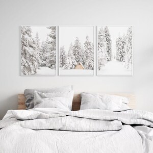 May include: A set of three vertical wall art prints in white frames, each featuring a black and white winter landscape of snow-covered pine trees. The center print includes a small, light-colored wooden cabin nestled among the snowy forest.