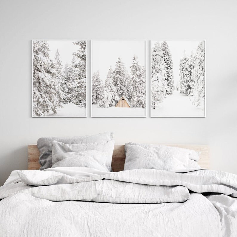 May include: A set of three vertical wall art prints in white frames, each featuring a black and white winter landscape of snow-covered pine trees. The center print includes a small, light-colored wooden cabin nestled among the snowy forest.